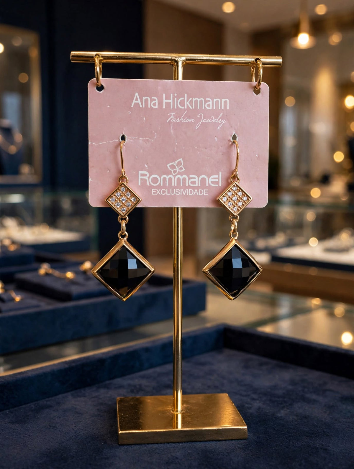 Par de brincos pendente ónix losango zircónias Ana Hickmann Rommanel em bandeja branca vitrine loja joalharia bokeh dourado