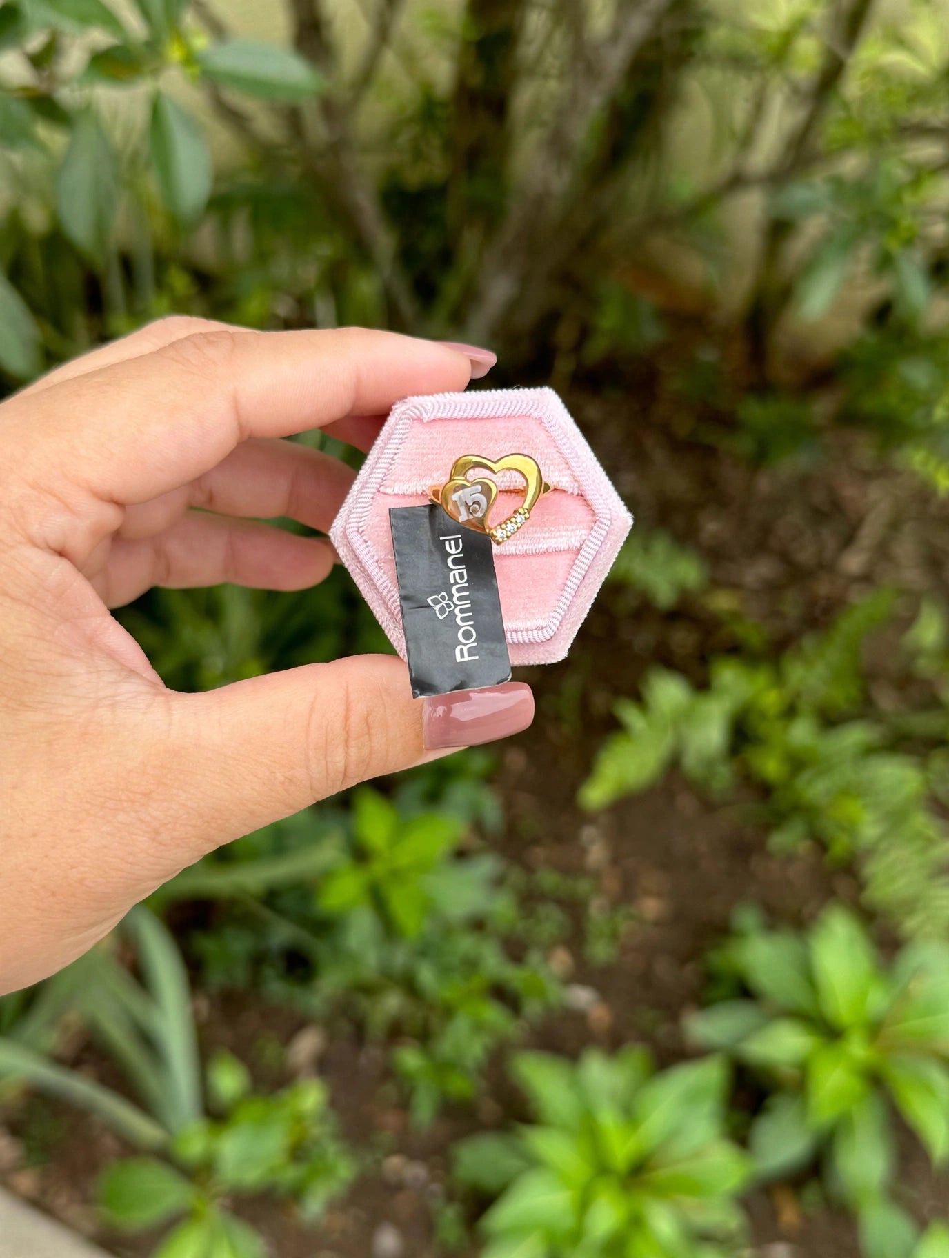 Anel 15 Anos Dois Corações Entrelançados Rommanel - na caixa rosa hexagonal com etiqueta Rommanel fundo jardim verde