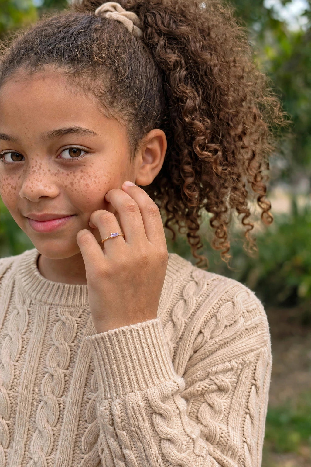 Anel Infantil Zircónia Roxa Baguette Rommanel - modelo criança com cabelo encaracolado a tocar no rosto fundo jardim verde